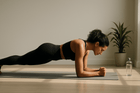 A woman in a black workout outfit performing a plank exercise on a yoga mat in a well-lit room, with a plant and a water bottle nearby.