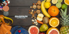 A colorful assortment of healthy fruits and nuts including oranges, bananas, walnuts, and kiwis, alongside fitness items like a measuring tape, a towel, and weights, showcasing the connection between nutrition and muscle building.