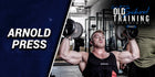 Image of a person performing the Arnold Press, a shoulder exercise involving dumbbells, demonstrating proper form with focused expression in a gym setting.