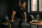 A focused young man performing a deadlift in a gym, showcasing strong arm muscles. In the foreground, a plate with healthy food including salmon, broccoli, and carrots alongside a water bottle, emphasizing a balanced diet for fitness.